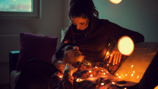 Une femme et un homme étendus ensemble sur un sofa et couverts de guirlandes de Noël lumineuses se regardent.