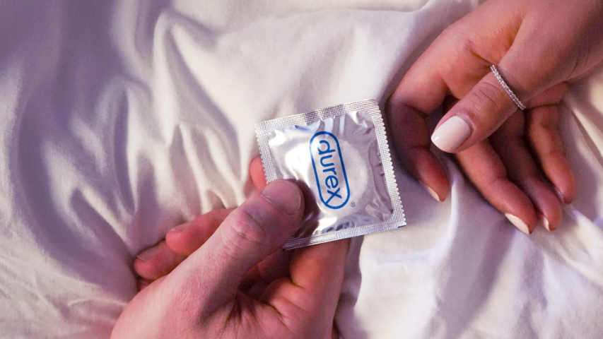 Two hands passing each other a silver-wrapped Durex condom in bed. 