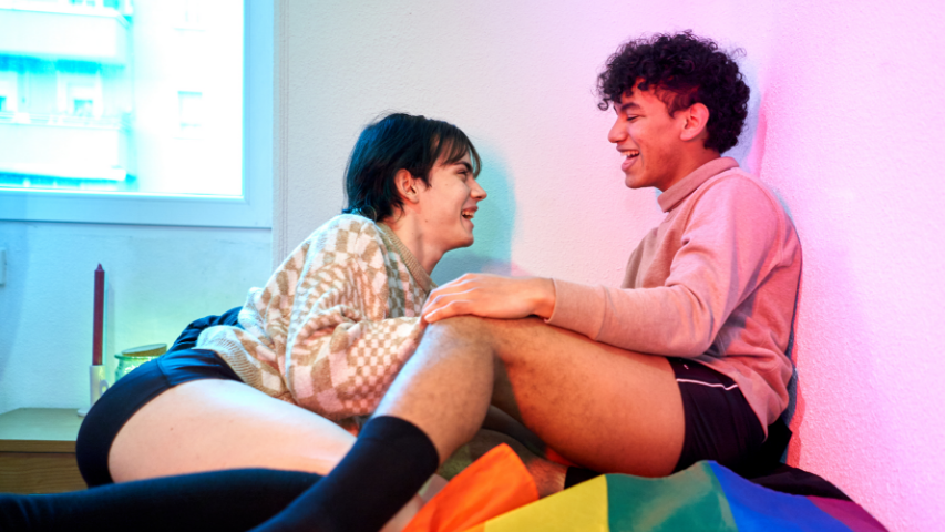 Two people having a spirited discussion about trans sex while sitting on top of a Pride flag in a well lit room.