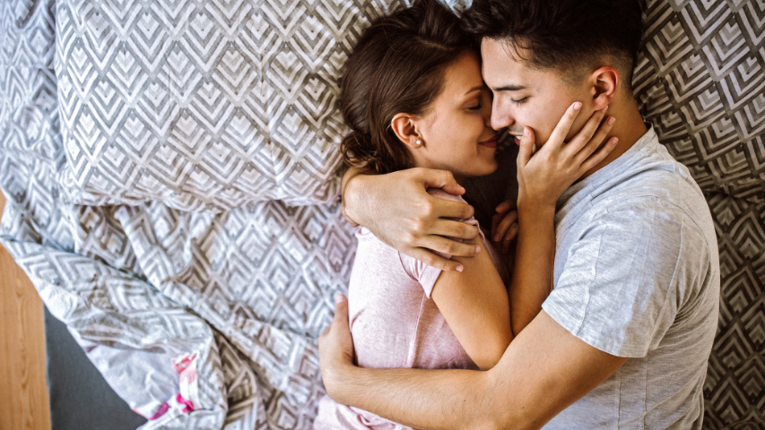 Two people holding each other tightly in bed on top of grey patterned pillowcases and bed sheets. 