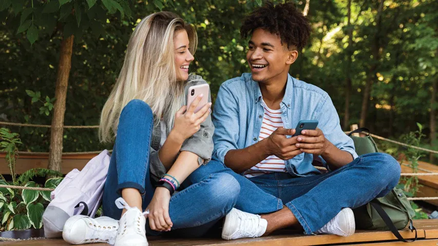 Two young people with backpacks sitting on a public bench looking at each other and laughing while holding out their cell phones.