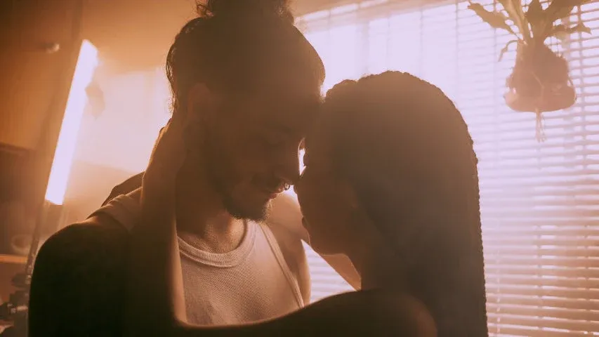 Couple leaning in to kiss one another in front of a sunlit blind.